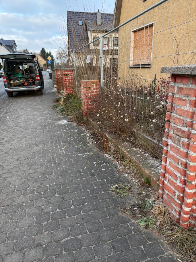 Ziegelmauer und Gehweg mit verwildertem Pflanzenbewuchs neben einem Lieferwagen.